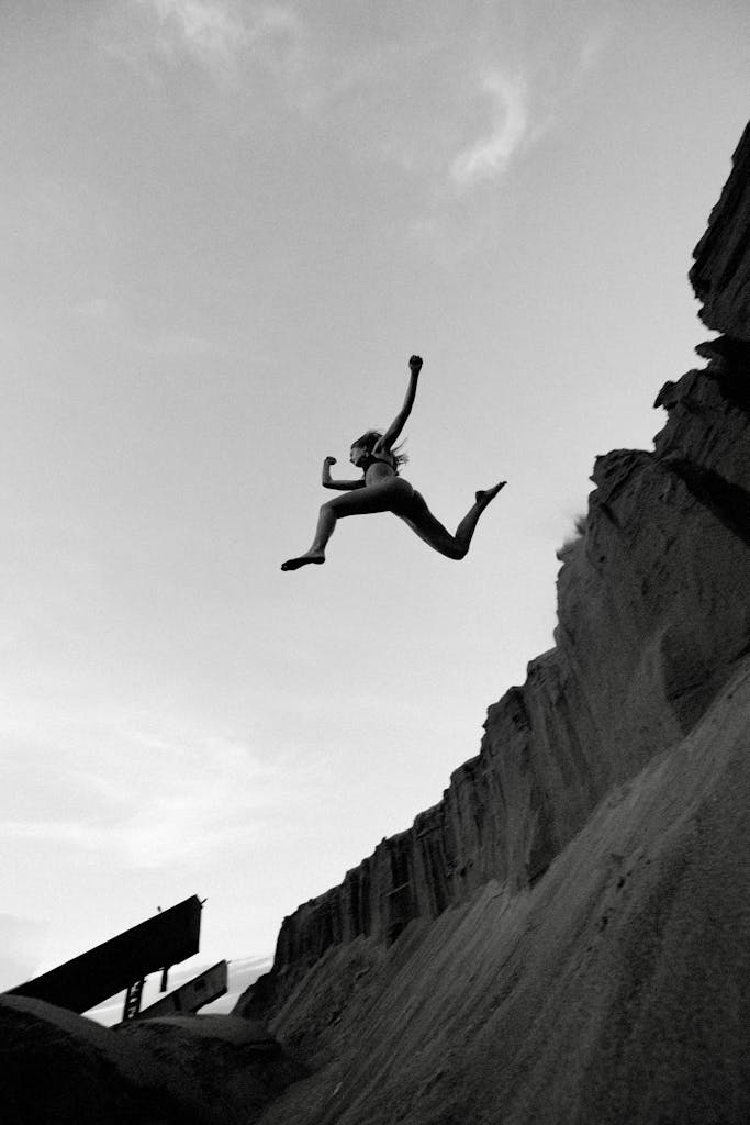 A person energetically leaps over dramatic cliff formations in black and white.