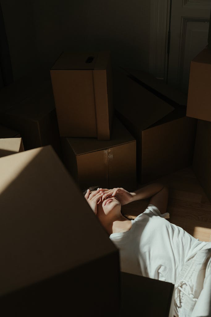 A woman lies on the floor surrounded by cardboard boxes in a dim sunlit room, expressing fatigue.