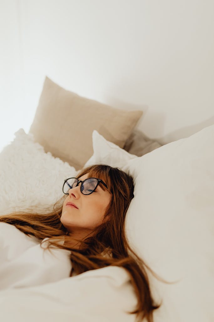A woman relaxes comfortably in bed with glasses on, surrounded by soft, fluffy pillows.