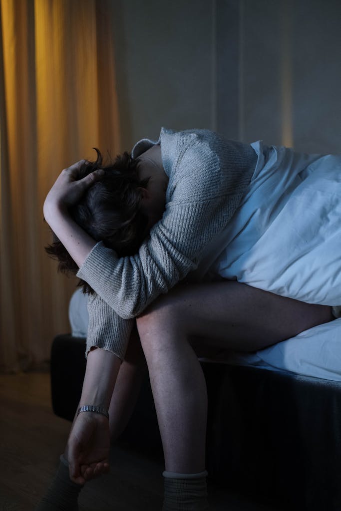 A woman sits on her bed, experiencing anxiety and insomnia in a dimly lit room.