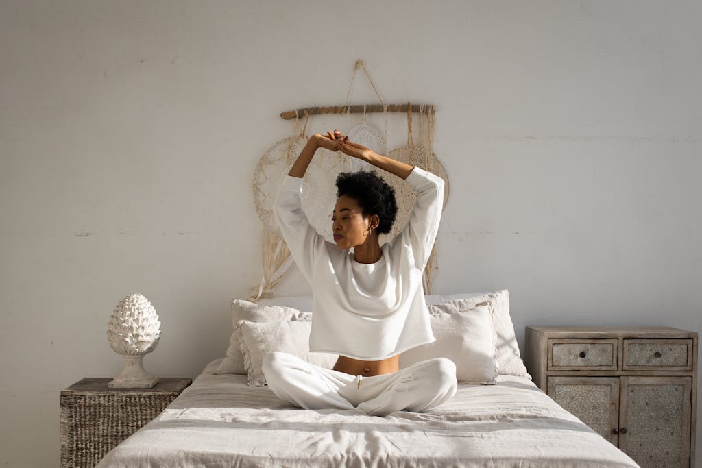 A woman sitting cross-legged and stretching in a tranquil bedroom setting, capturing a peaceful morning moment.
