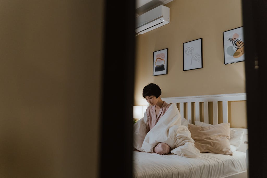Asian woman sitting on bed, wrapped in blanket, indoors depicting feelings of loneliness.