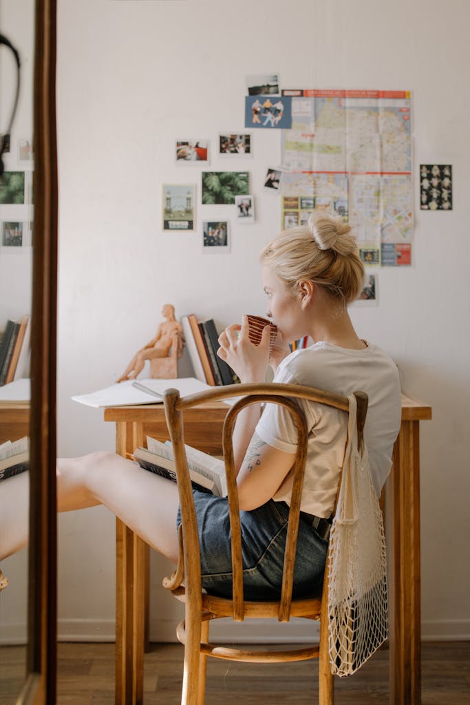 Back view of a blonde woman sipping tea in a cozy dorm setting with photos and books on the wall.